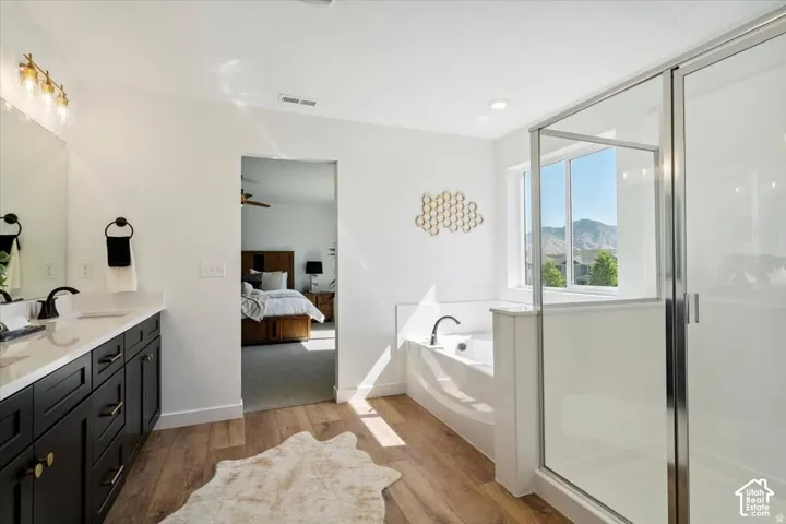 Dual vanity with dark cabinetry, wood-finish flooring, built-in bathtub, glass-enclosed shower, and large window