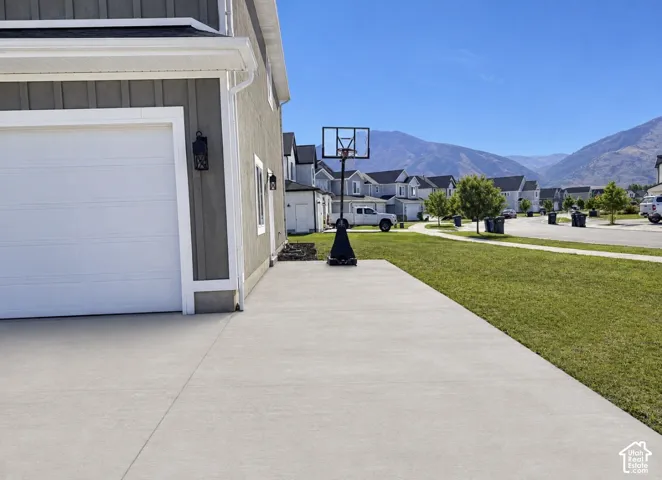 Third Car/ RV Pad: Concrete driveway, attached garage, exterior wall lantern, mountain backdrop, and green lawn