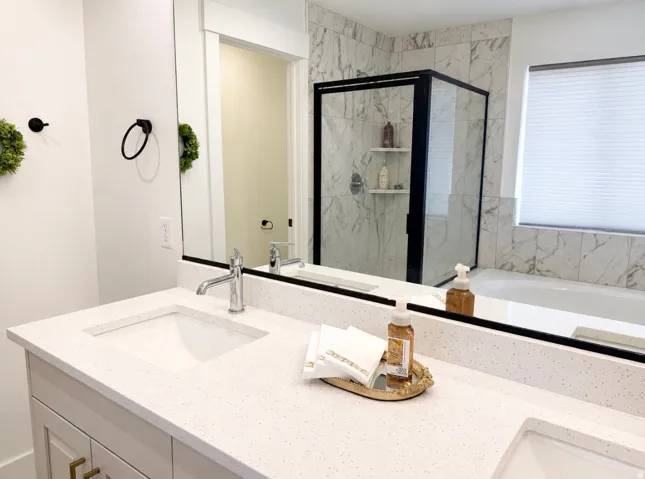 Dual undermount sinks with chrome faucets, quartz countertop, large vanity mirror, walk-in shower with black-framed glass enclosure, and a built-in soaking tub