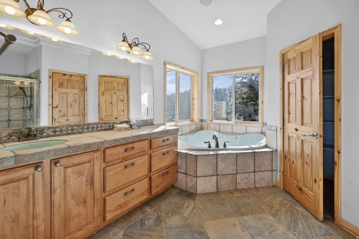 Dual vanity with stone countertop and integrated sinks, corner jetted tub with tiled surround, and wood-finish cabinetry