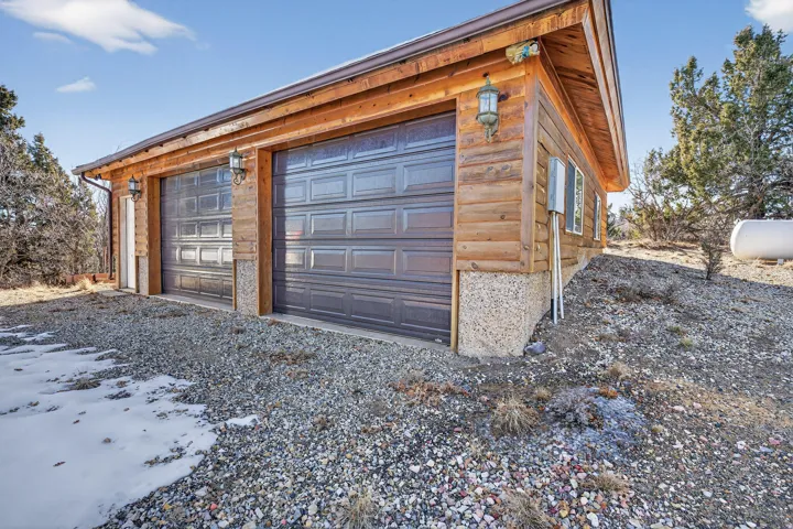 Wood-clad garage structure with two panel-style garage doors, exterior sconce lighting, and a gravel surface