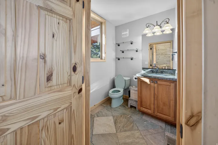 Wood panel door opening to a bathroom featuring stone tile flooring, a wood vanity with a stone countertop, a wall-mounted mirror, and a window with wood trim