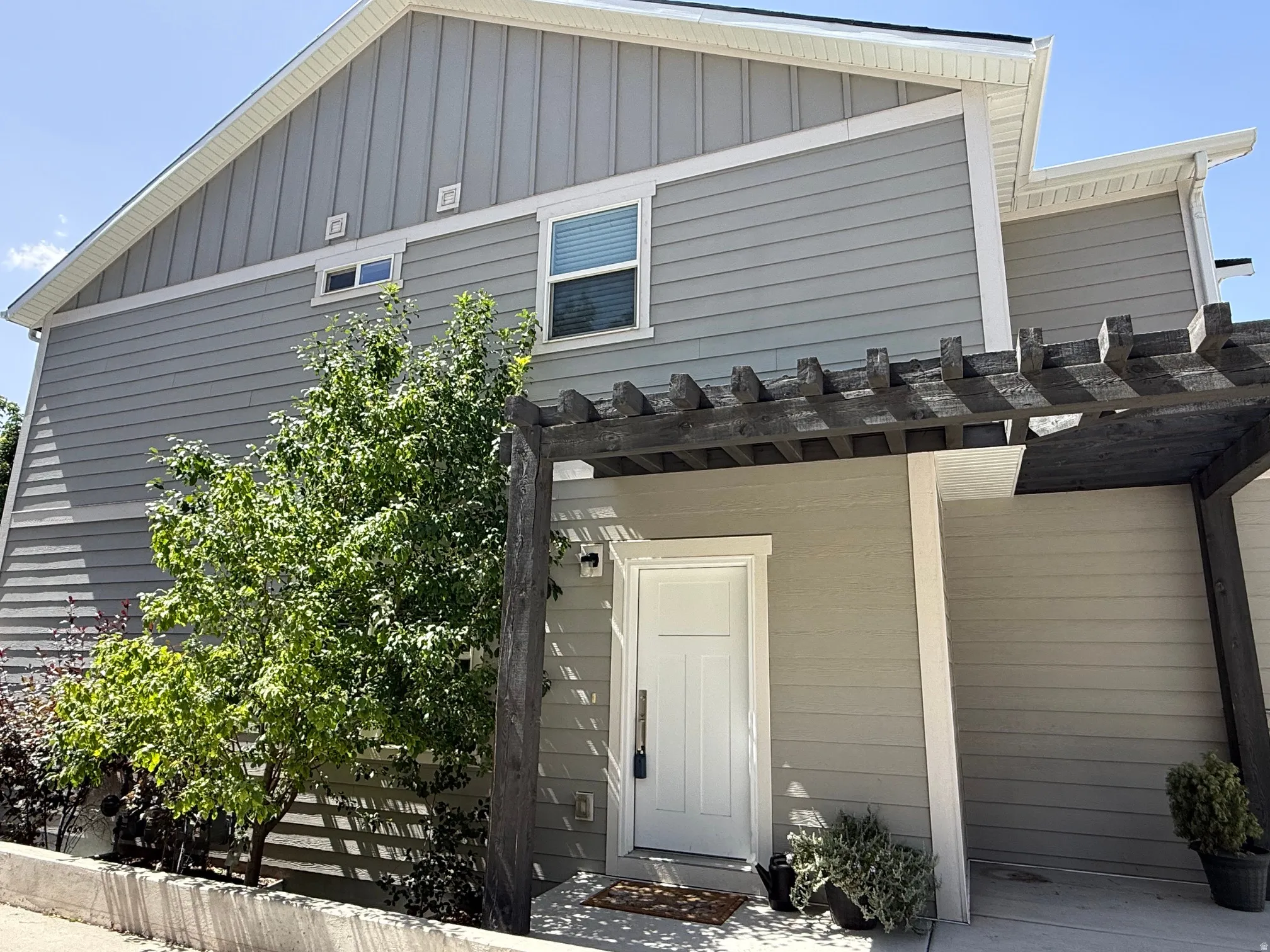 Exterior featuring light gray horizontal siding, vertical gable siding, a white entry door, and a dark wood pergola