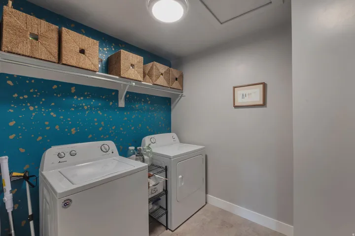 Dedicated laundry room featuring side-by-side white appliances, abundant storage with woven baskets on floating shelving, decorative blue accent wall with gold leaf detailing, recessed ceiling lighting, and neutral wall finishes