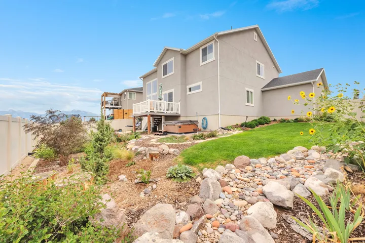 Multi-story stucco exterior with a composite deck, a lower-level hardscaping feature, a manicured lawn, and natural landscaping