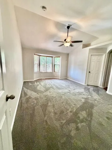 Spacious room featuring vaulted ceilings, a ceiling fan with integrated lighting, a bay window, and soft carpet flooring