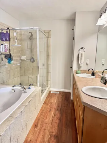 Dual vanity with integrated sinks, wood-finish flooring, tiled soaking tub, and a glass-enclosed shower with tiled surround
