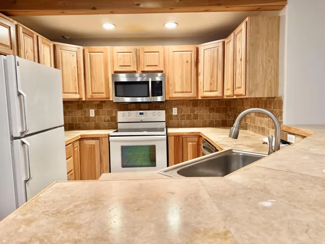 Light wood cabinetry with recessed panel doors, tile countertops, stainless steel sink with gooseneck faucet, full tile backsplash, and recessed lighting