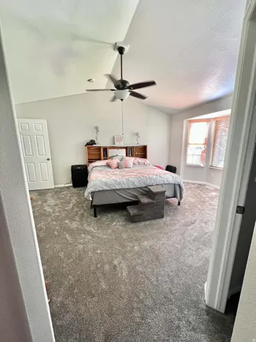 Vaulted ceiling, ceiling fan with light fixture, bay window, two-panel interior door, and grey patterned carpeting