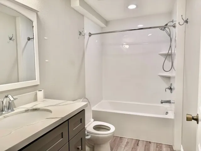 Bathroom featuring a single vanity with stone-finish countertop, wood-finish flooring, a white framed mirror, and a shower-tub combination with built-in shelving