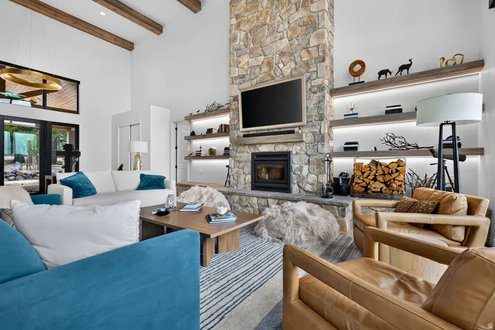 Vaulted ceiling with exposed wood beams, stone fireplace, built-in shelving with integrated lighting, wood-finish flooring, and large glass-paned doors