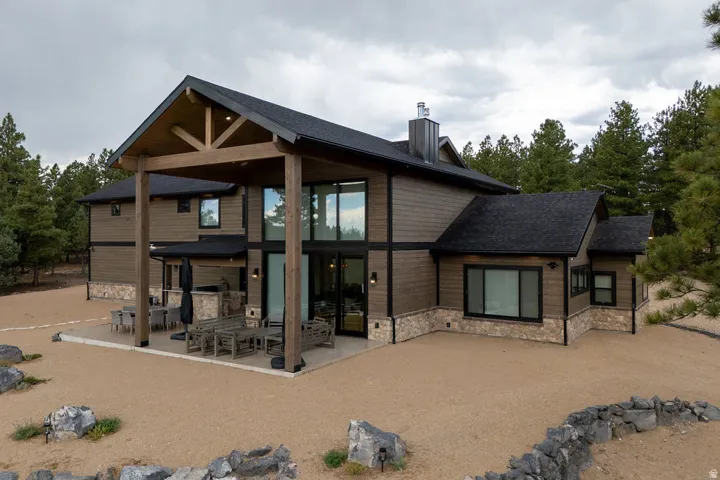 Contemporary residence featuring wood siding, stone accents, a covered patio with exposed beam detailing, expansive windows, and a dark shingle roof