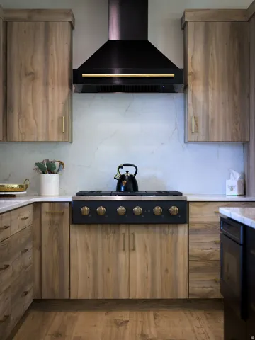 Wood-finish cabinetry with brass hardware, a black range hood, a countertop range, a light-toned backsplash, and a light-toned countertop