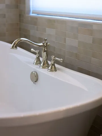 Freestanding bathtub with polished nickel fixtures, complemented by a tiled wall and a window with a roller shade