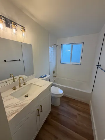Modern bathroom featuring a marble-look vanity countertop with dual brass fixtures, white cabinetry with ample storage, frameless glass shower enclosure, soaking tub, and wood-finish flooring throughout