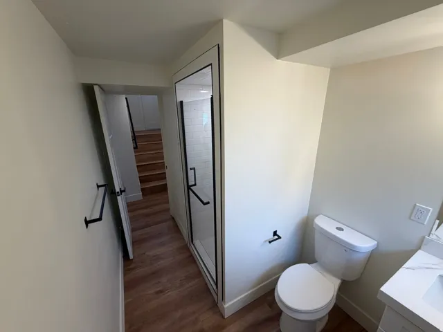 Glass-enclosed shower with black trim, tile shower surround, wood-finish flooring, white toilet, and white vanity with an integrated sink