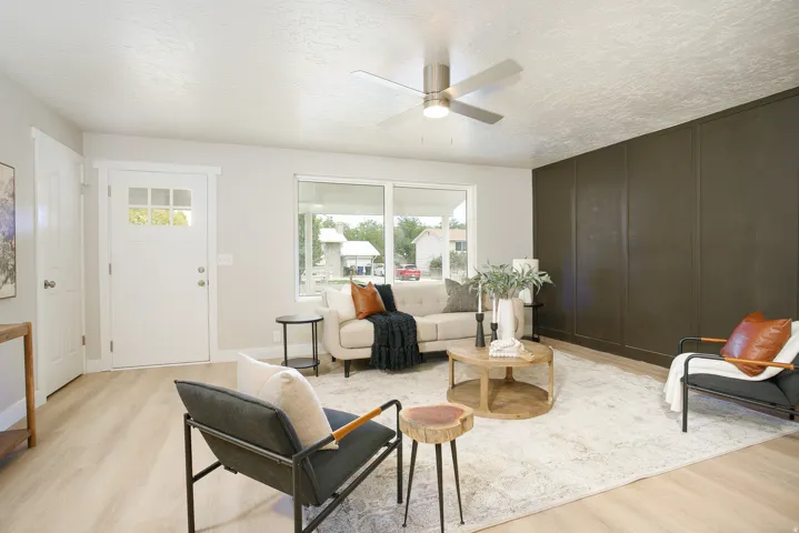 Wood-finish flooring, accent wall with panel detailing, large picture window, ceiling fan with integrated lighting, and a front entry door with glass inserts