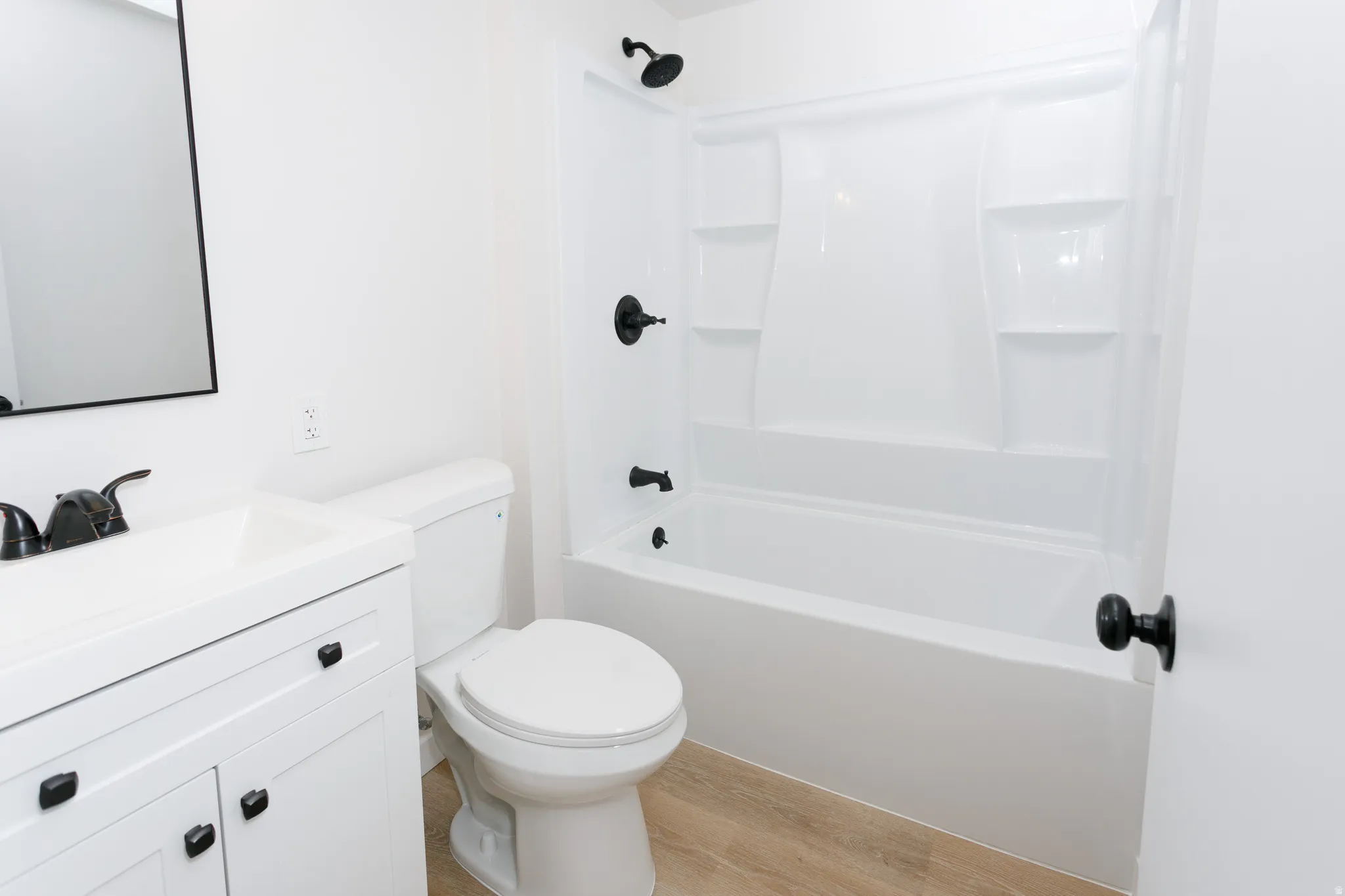 White vanity with integrated sink, black hardware, white toilet, white tub-shower combination, and wood-finish flooring