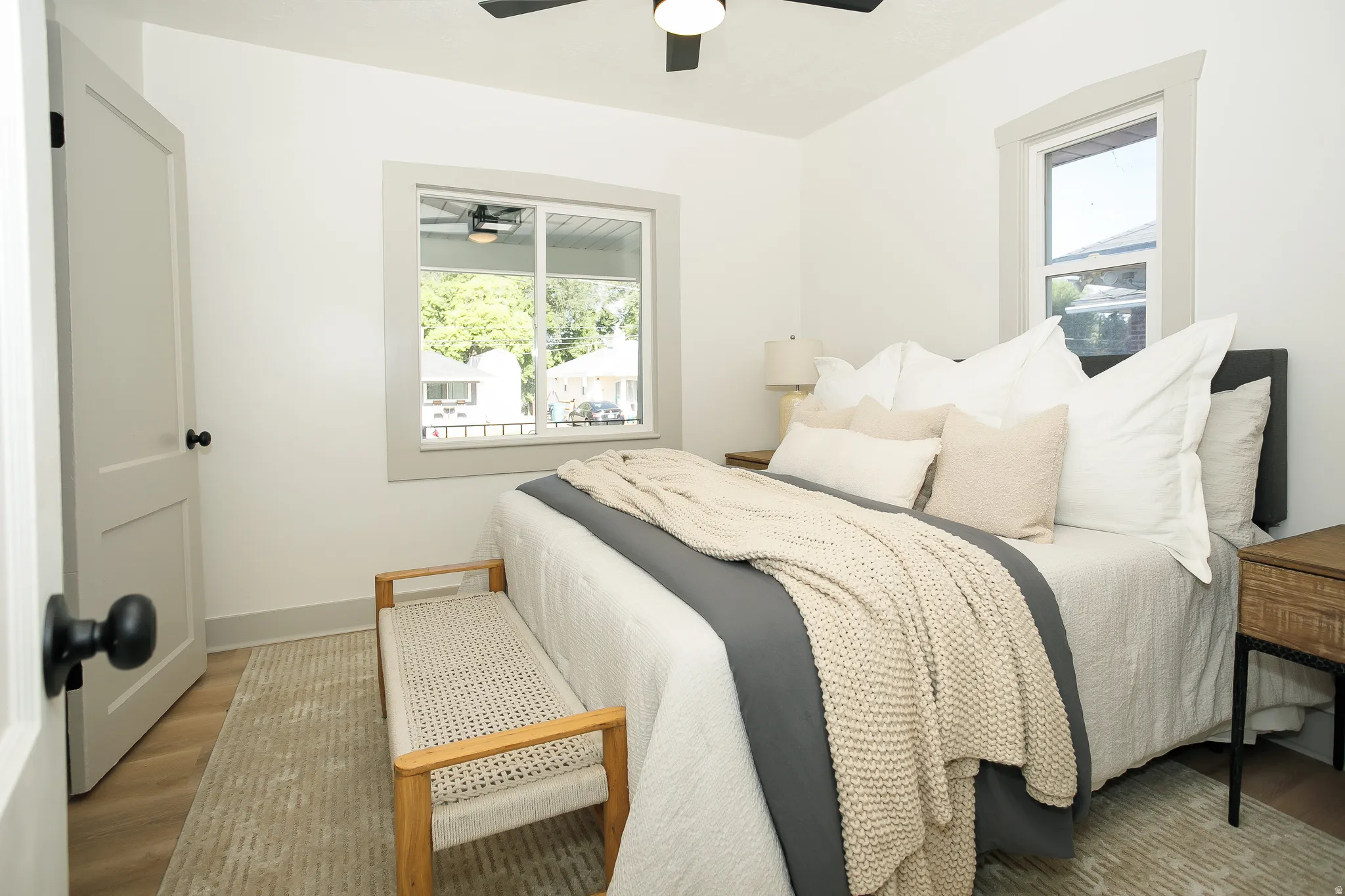 White walls, light wood-finish flooring, two windows with gray trim, a ceiling fan, and a white interior door with black hardware