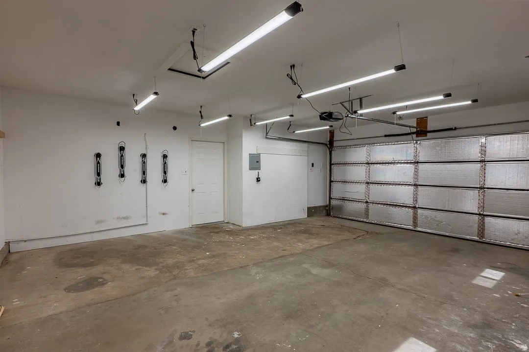 Concrete flooring, multiple overhead LED light fixtures, a garage door with an opener, a white interior door, and a wall-mounted electrical panel