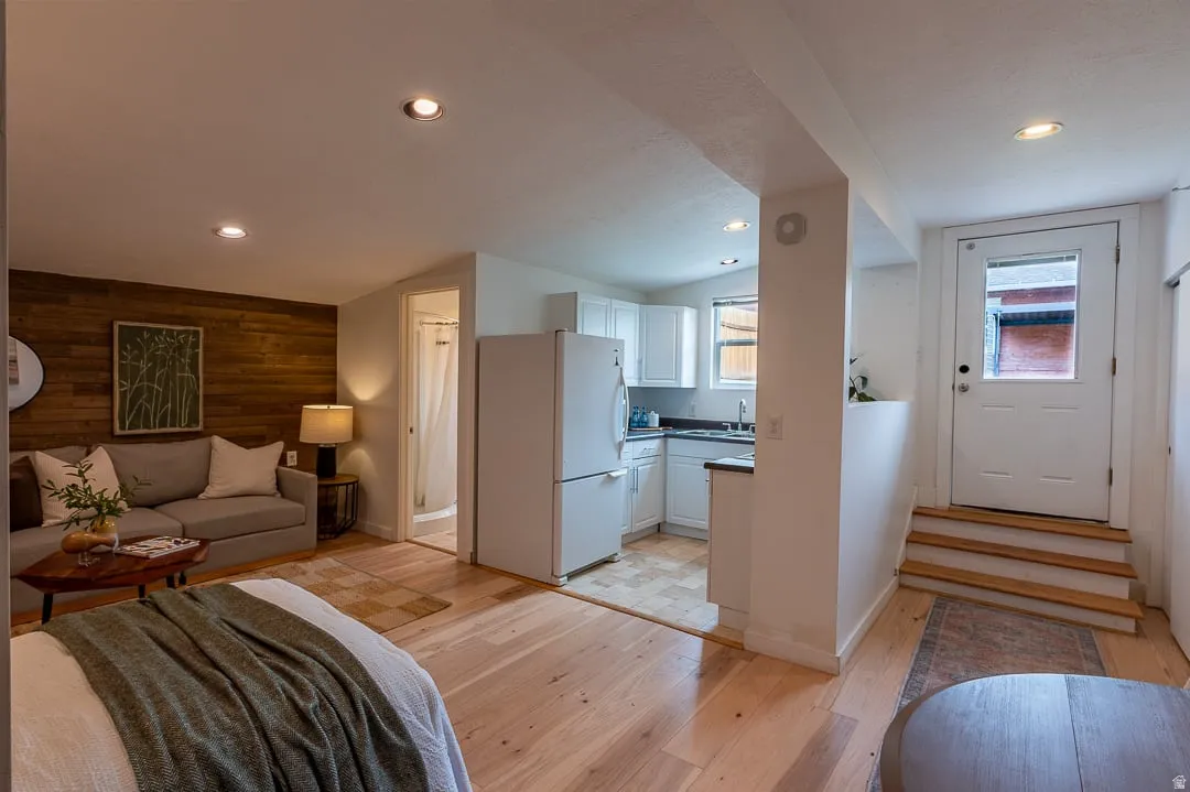 Open-concept studio featuring warm wood-finish flooring throughout, a wood-accent accent wall, and a kitchenette with white cabinetry and stainless steel appliances