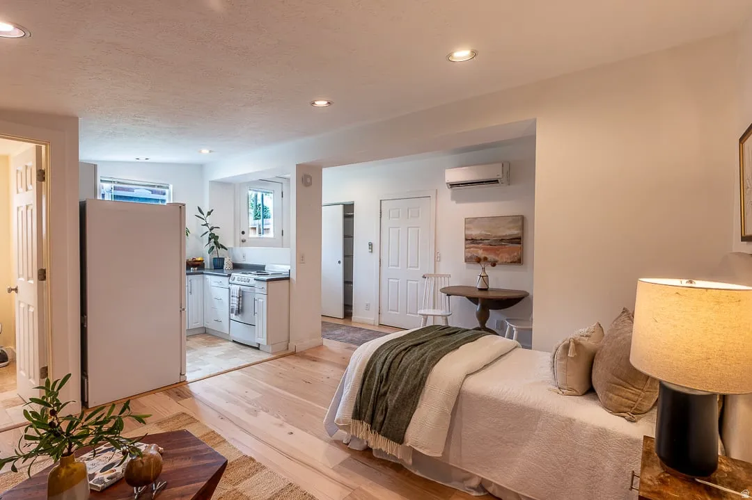 Efficient studio layout featuring wood-finish flooring throughout, a compact kitchenette with white cabinetry and standard appliances, a bed area with built-in bedside lighting, and recessed ceiling fixtures