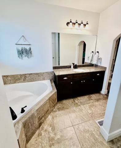Corner soaking tub with tiled surround, dark wood-finish vanity with granite countertop, undermount sink, and tiled flooring