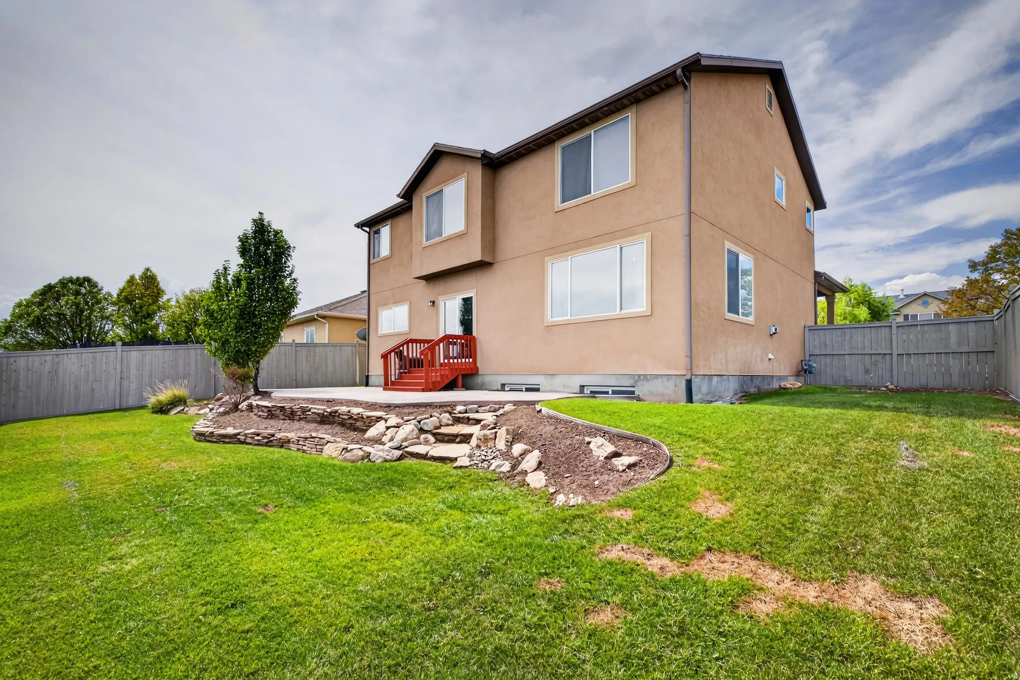 Expansive backyard featuring a manicured lawn, tiered concrete patio, and a red wood-finish deck with stairs