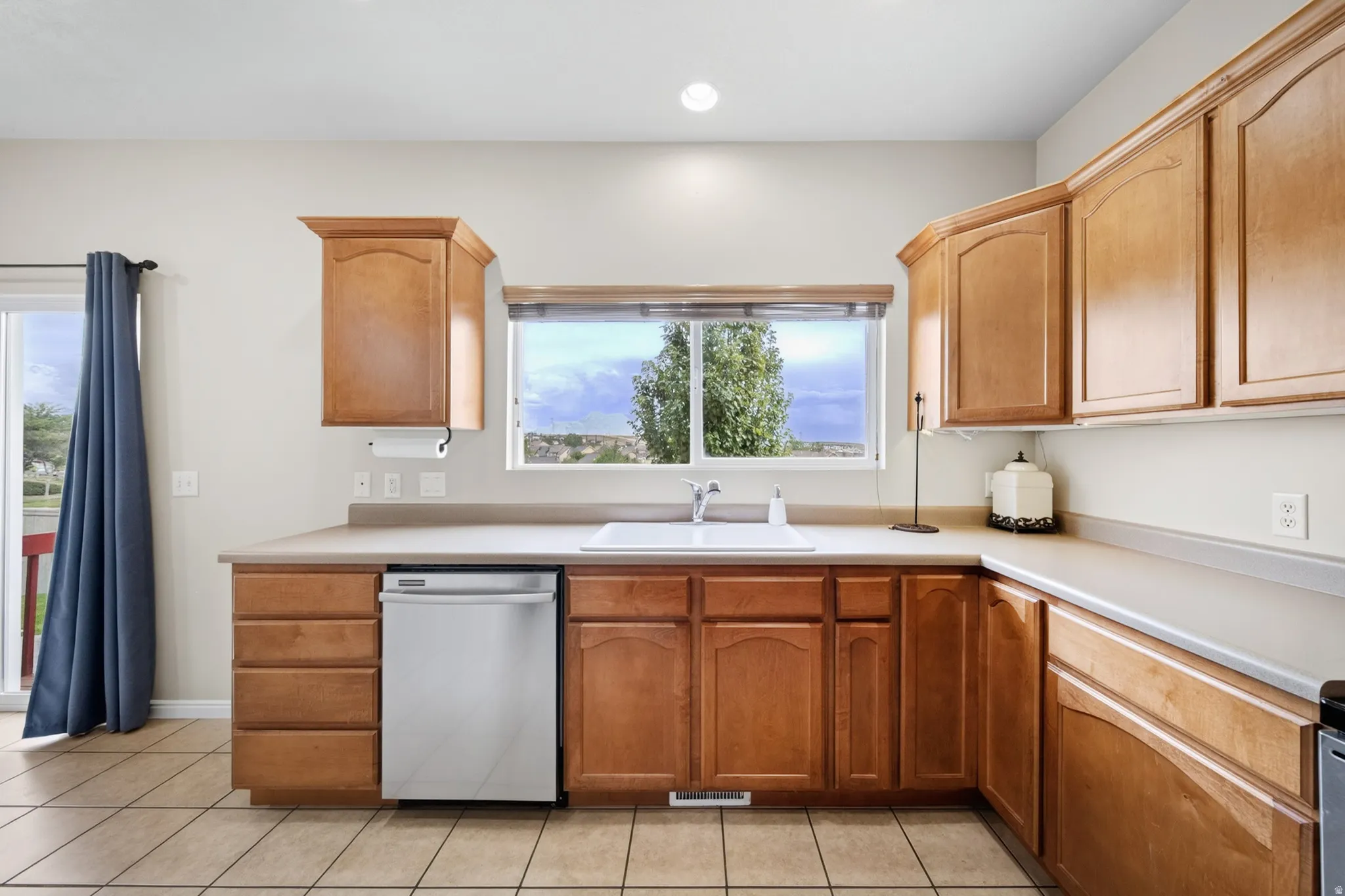 Wood-finish cabinetry, tile flooring, stainless steel dishwasher, white sink, and light-toned countertops