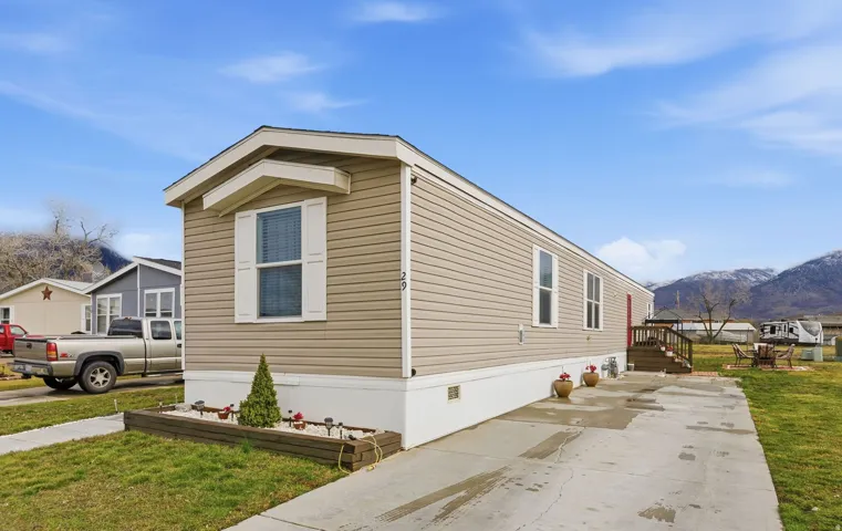 Beige vinyl siding exterior with white window shutters, a front entry deck with wood railing, a concrete driveway, and landscaped front planting bed