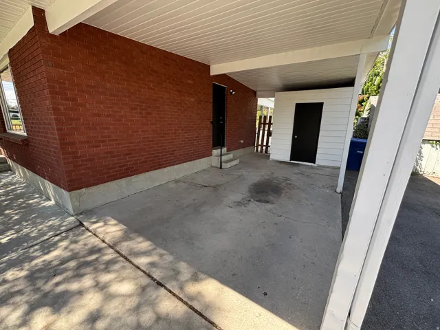 Covered concrete carport featuring a storage shed with a black door, exterior brick siding, and a wooden privacy fence