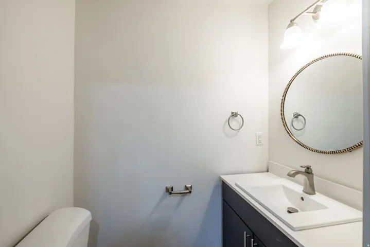 White vanity with integrated rectangular sink, brushed nickel faucet, round mirror with textured frame, and a two-light vanity fixture