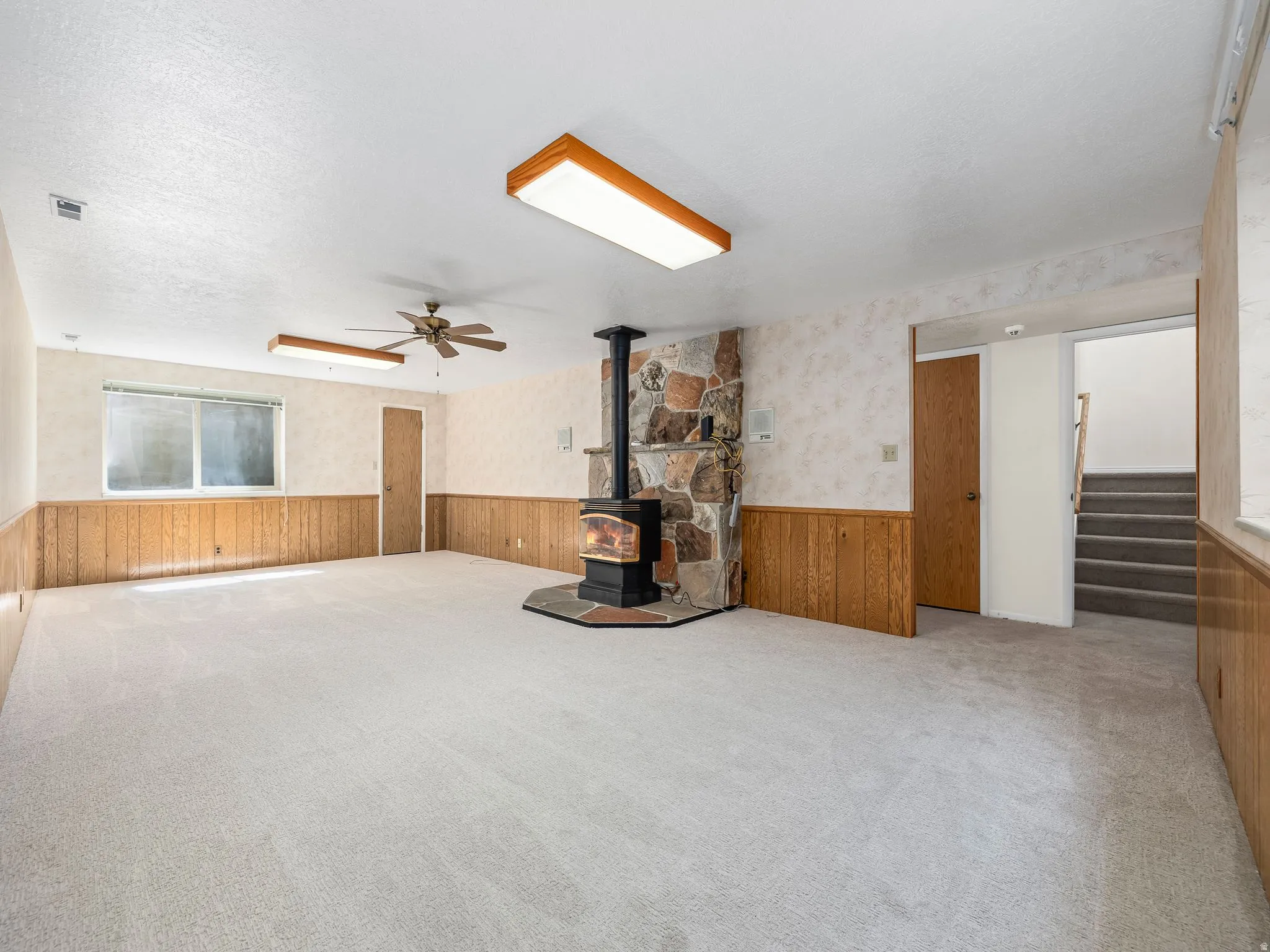Spacious room featuring a stone-clad wood-burning stove, wall-mounted light fixtures, a ceiling fan, and carpeted flooring