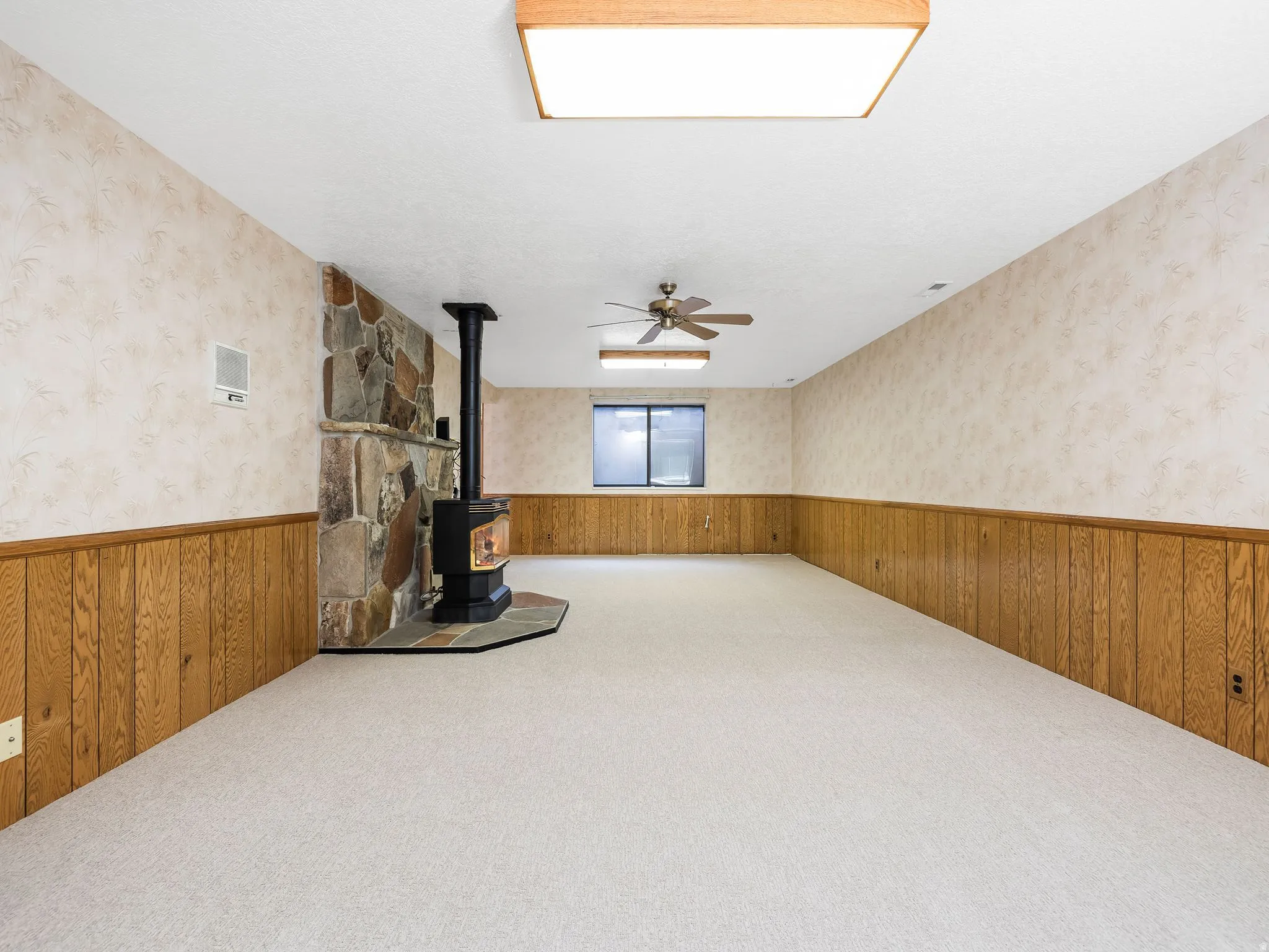 Spacious room featuring a stone-clad wood-burning stove, light-toned carpeting, wood paneling, and two ceiling light fixtures