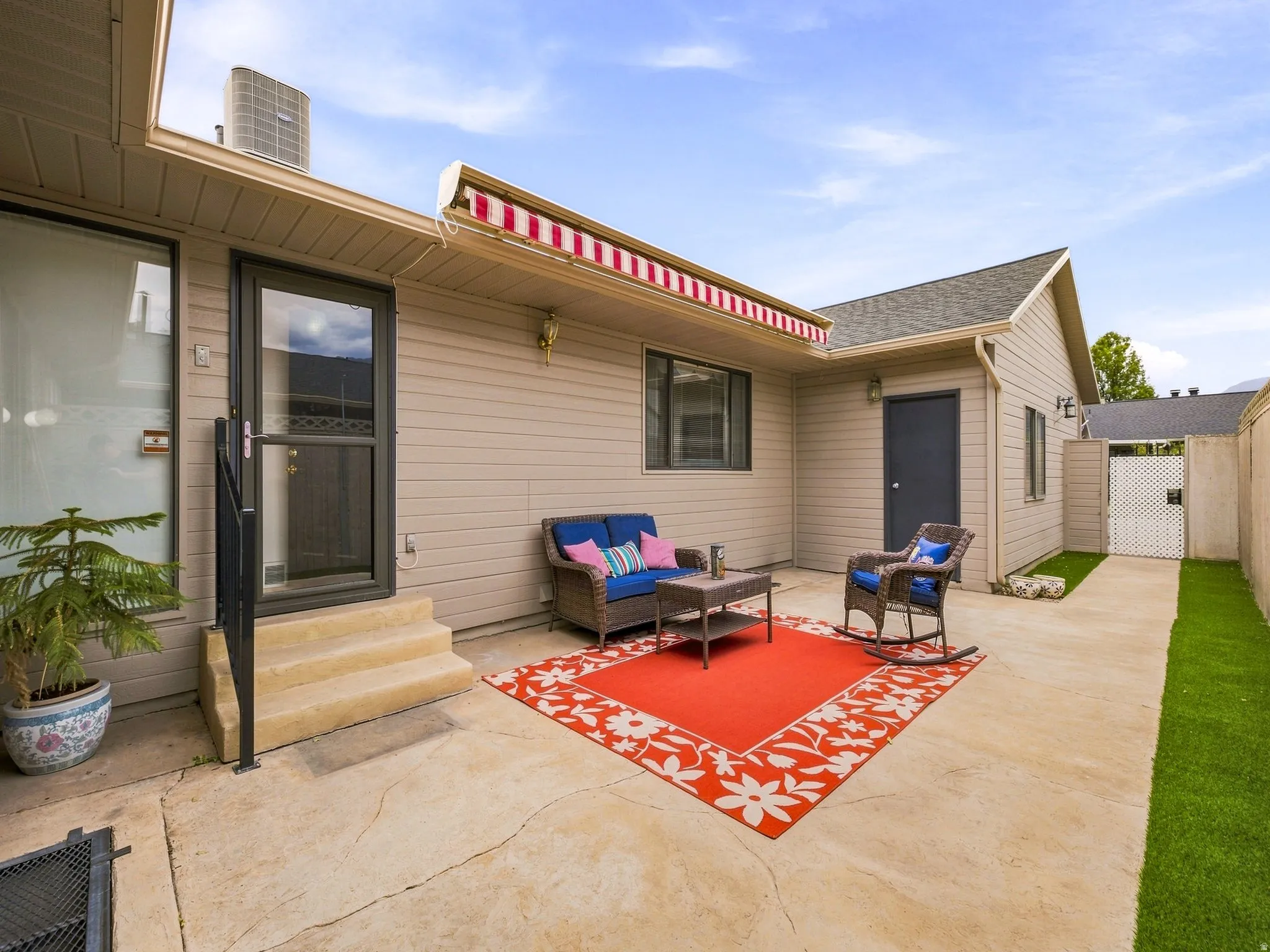 Exterior patio with a retractable striped awning, concrete hardscaping, and a fully enclosed side yard with artificial turf