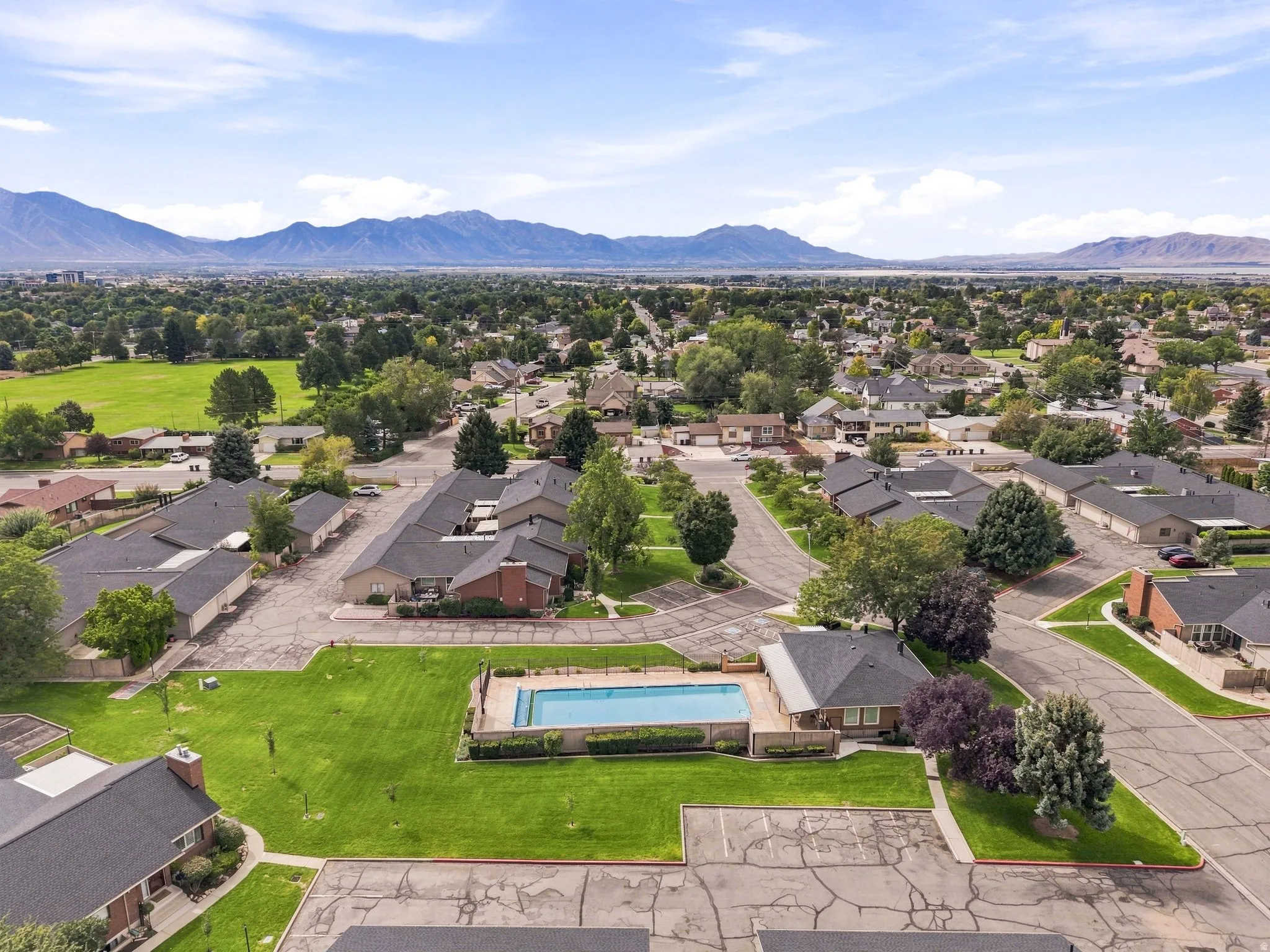 Community swimming pool with surrounding green space, residential buildings with dark shingle roofs, paved roadways, and distant mountains