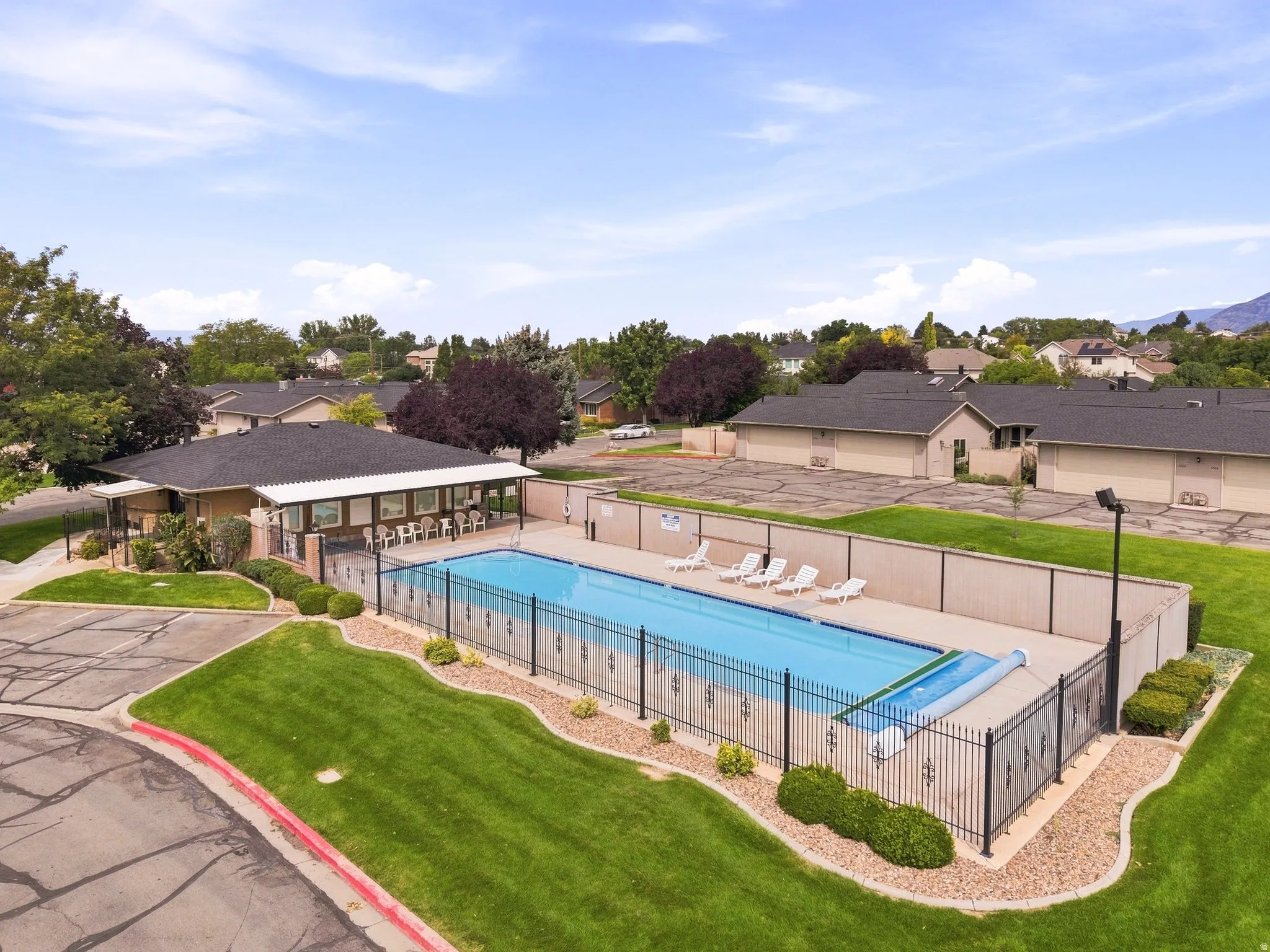 Outdoor swimming pool with surrounding black wrought iron fencing, an adjacent clubhouse featuring covered patio seating, manicured landscaping, and a paved parking area