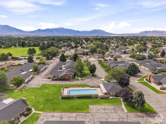 Community swimming pool with surrounding green space, residential buildings with dark shingle roofs, paved roadways, and distant mountains