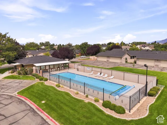 Outdoor swimming pool with surrounding black wrought iron fencing, an adjacent clubhouse featuring covered patio seating, manicured landscaping, and a paved parking area