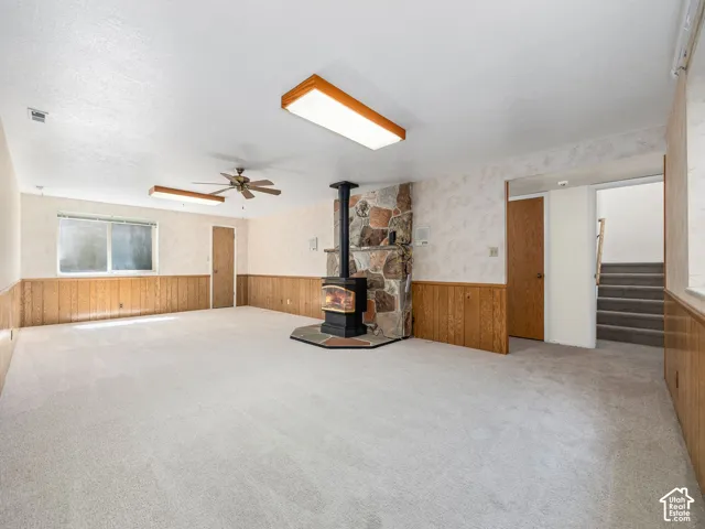 Spacious room featuring a stone-clad wood-burning stove, wall-mounted light fixtures, a ceiling fan, and carpeted flooring