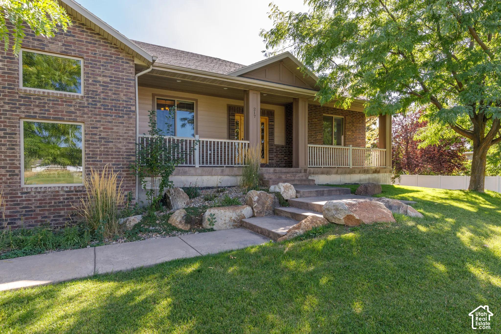 Brick exterior with multiple windows, covered porch with columns, a white railing, stone landscaping, and mature trees