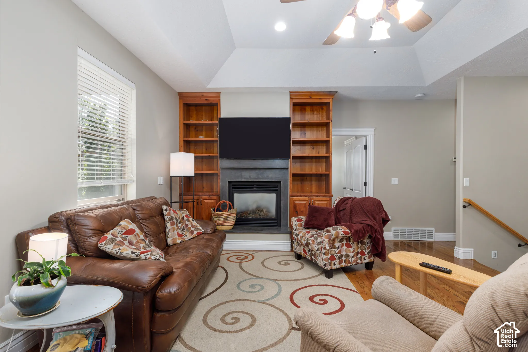 Vaulted ceiling with a ceiling fan, gas fireplace with stone surround, wood-finish built-in shelving, and wood-finish flooring