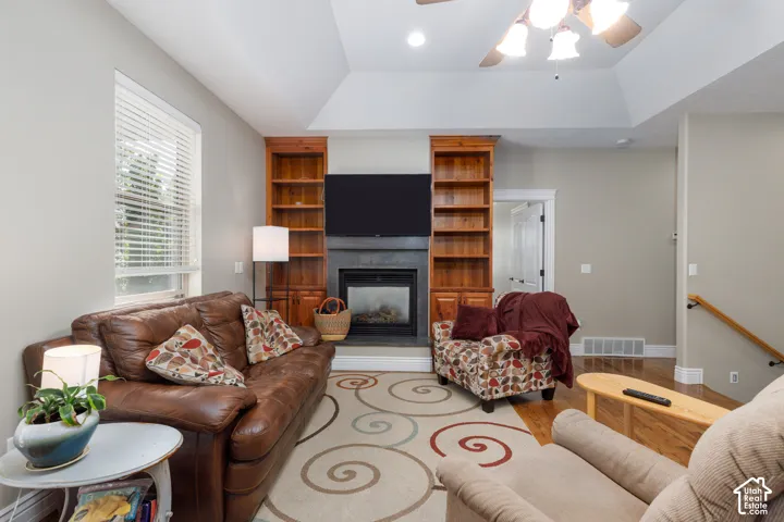 Vaulted ceiling with a ceiling fan, gas fireplace with stone surround, wood-finish built-in shelving, and wood-finish flooring