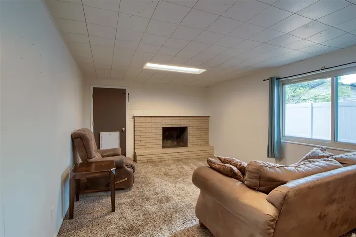 Plush carpet flooring, brick fireplace, drop ceiling with recessed lighting, and a large window