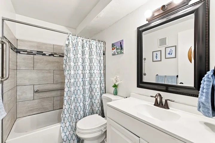 Bathtub with light-toned tiled surround, vanity with white countertop and integrated sink, bronze-finish faucet, and framed mirror with integrated lighting. Updated in 2021