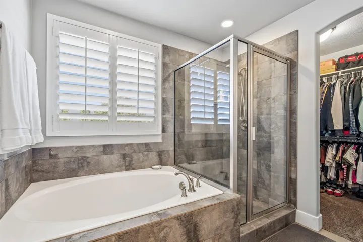 Built-in soaking tub with chrome fixtures, glass-enclosed shower with tile surround, and window with plantation shutters