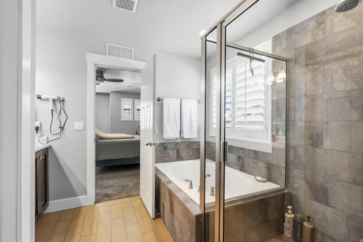Built-in tub with tile surround, glass-enclosed shower with tile walls, wood-finish flooring, and window with plantation shutters
