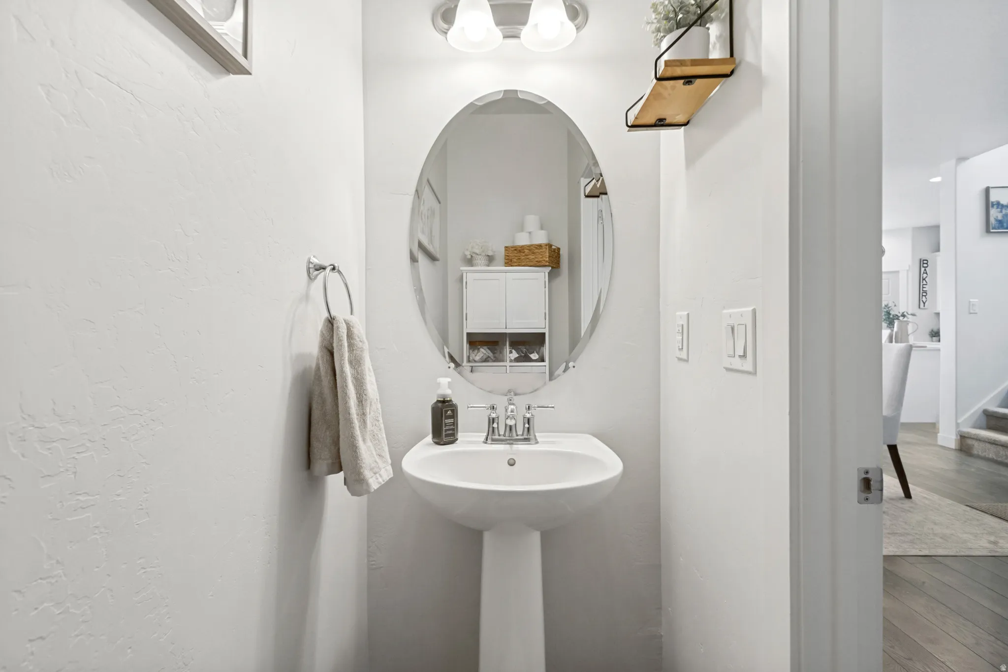 Elegant powder room featuring a pedestal sink with a round vessel basin, oval framed mirror with integrated shelf storage, white textured walls, and polished chrome fixtures