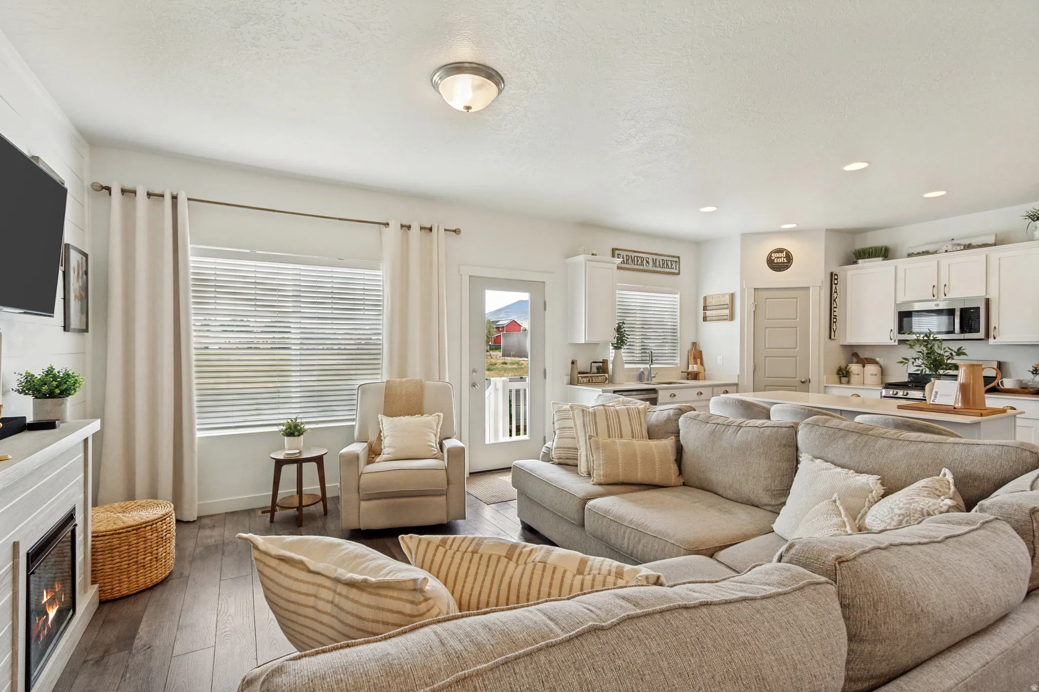 Open-concept living area featuring neutral upholstered seating, wood-finish flooring throughout, recessed ceiling lighting, and large windows with operable shutters and curtain hardware