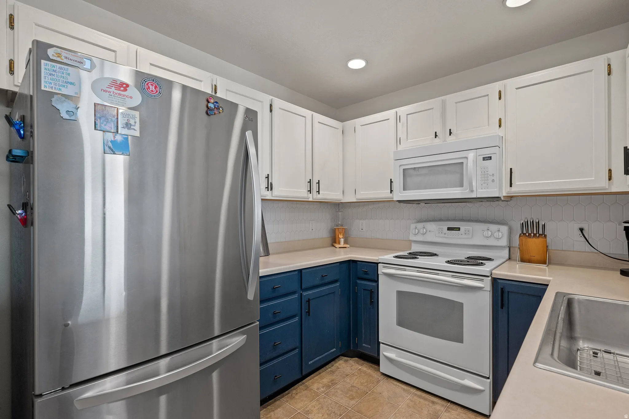 Stainless steel refrigerator, white upper cabinetry, blue lower cabinetry, light-toned countertops, and a white electric range with an over-the-range microwave